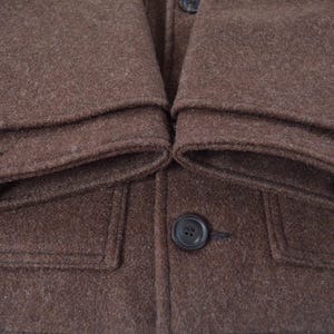 May include: Close-up of a brown wool coat. The coat has a buttoned front, two front pockets, and buttoned cuffs. The fabric has a textured appearance.
