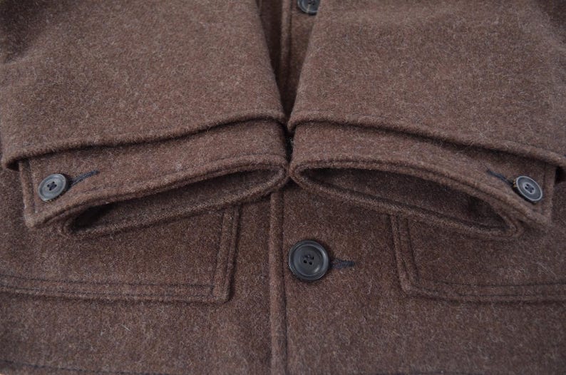 May include: Close-up of a brown wool coat. The coat has a buttoned front, two front pockets, and buttoned cuffs. The fabric has a textured appearance.