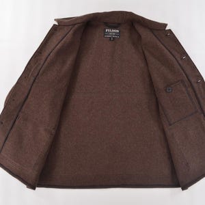 May include: Open brown jacket with a button closure and two front pockets. The jacket has a collar and a label that reads "FILSON". The jacket is made of a thick, textured material.