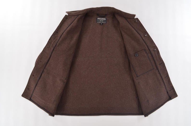May include: Open brown jacket with a button closure and two front pockets. The jacket has a collar and a label that reads "FILSON". The jacket is made of a thick, textured material.
