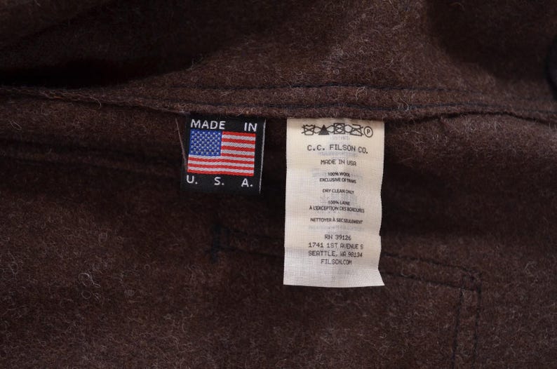 May include: Close-up of a brown wool garment with two tags. One tag displays the American flag and reads "MADE IN U.S.A.". The other tag has text including "C.C. FILSON CO." and care instructions.