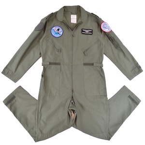 May include: An olive green military-style jumpsuit with a zip closure, pockets, and patches on the chest and sleeves. The jumpsuit features long sleeves and a collar. Patches include text and graphic designs. The jumpsuit is made of a durable fabric.