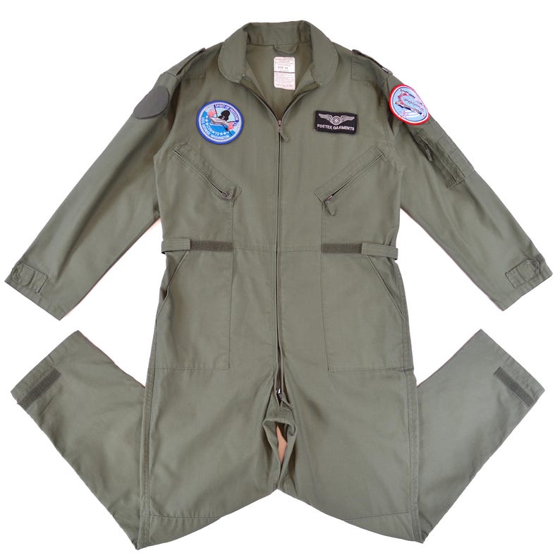 May include: An olive green military-style jumpsuit with a zip closure, pockets, and patches on the chest and sleeves. The jumpsuit features long sleeves and a collar. Patches include text and graphic designs. The jumpsuit is made of a durable fabric.