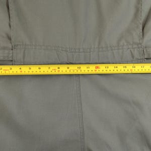 May include: Close-up of a sage green jumpsuit with a yellow measuring tape laid across the waist. The tape shows a measurement of approximately 46 cm. The jumpsuit has a belt and a seam down the front.