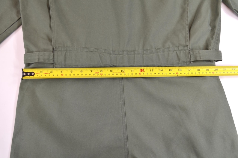 May include: Close-up of a sage green jumpsuit with a yellow measuring tape laid across the waist. The tape shows a measurement of approximately 46 cm. The jumpsuit has a belt and a seam down the front.