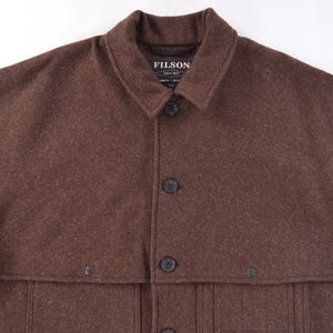 May include: A brown wool coat with a collar and button closure. The coat has a cape-like design over the shoulders and a label that reads "FILSON". The coat has multiple buttons and pockets.