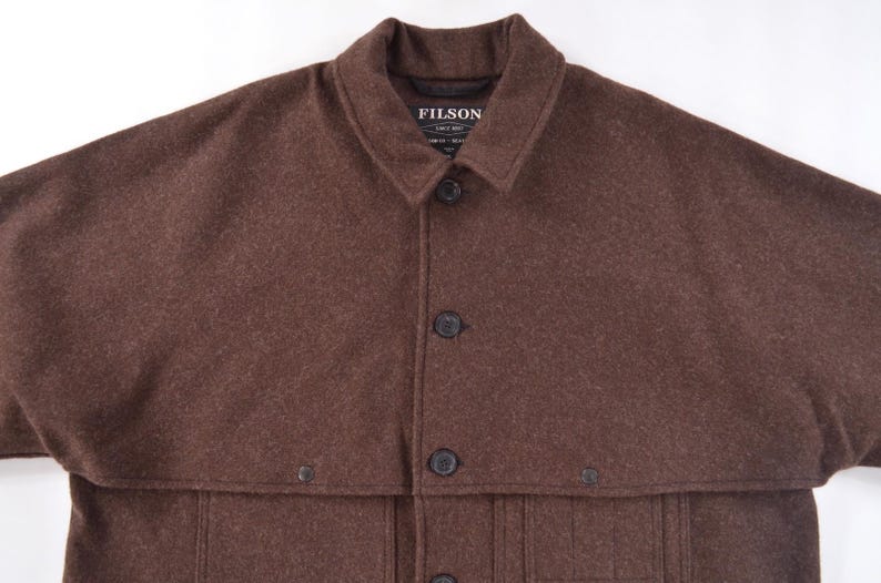 May include: A brown wool coat with a collar and button closure. The coat has a cape-like design over the shoulders and a label that reads "FILSON". The coat has multiple buttons and pockets.