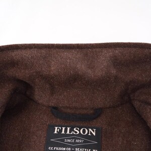 May include: A close-up shot of a brown wool coat collar. The coat is a dark brown shade with a textured finish. A black label displaying the brand name "FILSON" is visible, along with the text "SINCE 1897" and the company's location.