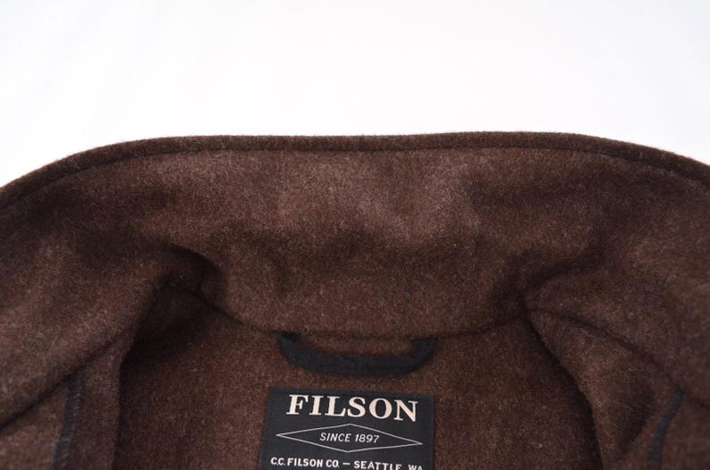 May include: A close-up shot of a brown wool coat collar. The coat is a dark brown shade with a textured finish. A black label displaying the brand name "FILSON" is visible, along with the text "SINCE 1897" and the company's location.