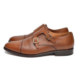 May include: A pair of brown leather double monk strap shoes. The shoes have a cap toe design and gold-coloured buckles. The shoes are set against a white background.