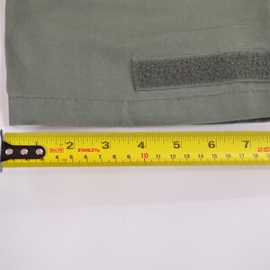 May include: A close-up of olive green fabric with a velcro patch. A yellow Stanley measuring tape is placed below the fabric, showing measurements up to 23 cm.