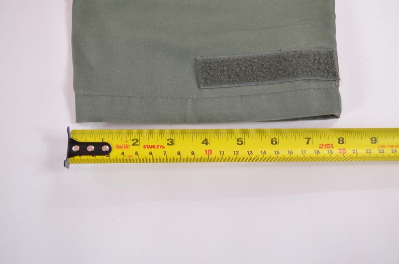 May include: A close-up of olive green fabric with a velcro patch. A yellow Stanley measuring tape is placed below the fabric, showing measurements up to 23 cm.
