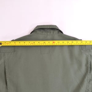 May include: An olive green garment with a collar and a yellow measuring tape laid across the back. The tape shows a measurement of approximately 58 cm across the shoulders. The garment has pockets and a button closure.