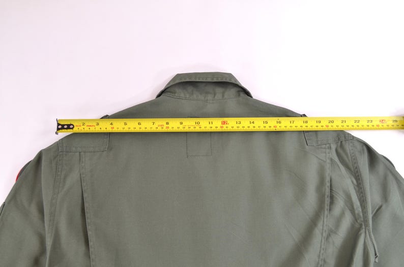 May include: An olive green garment with a collar and a yellow measuring tape laid across the back. The tape shows a measurement of approximately 58 cm across the shoulders. The garment has pockets and a button closure.