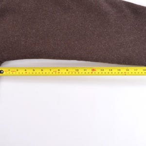 May include: A brown wool coat sleeve with a buttoned cuff. A yellow tape measure shows the sleeve's length. The sleeve measures approximately 56 cm from cuff to shoulder.