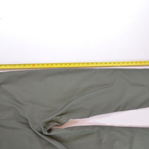 May include: Olive green trousers with a yellow measuring tape laid across the fabric. The trousers are laid out on a white surface. The tape measure shows the length of the trousers.