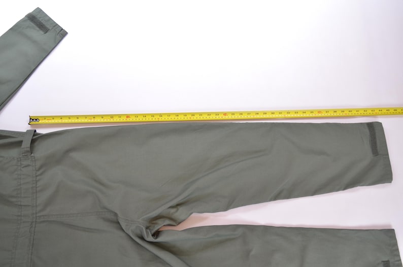 May include: Olive green trousers with a yellow measuring tape laid across the fabric. The trousers are laid out on a white surface. The tape measure shows the length of the trousers.