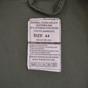 May include: Close-up of a green garment with a label. The label states "COVERALL, FLYERS CWU-27/P" and includes size "44". The fabric is 35% cotton and 65% polyester. Washing instructions are also printed on the label.