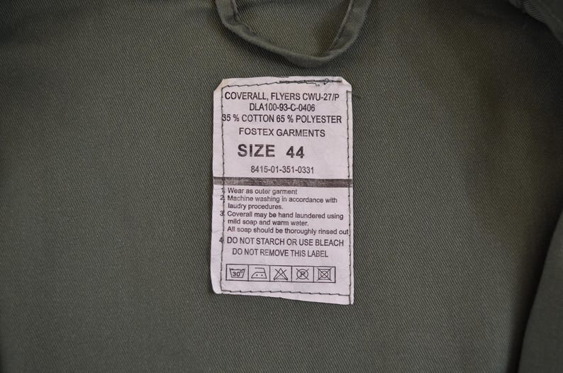 May include: Close-up of a green garment with a label. The label states "COVERALL, FLYERS CWU-27/P" and includes size "44". The fabric is 35% cotton and 65% polyester. Washing instructions are also printed on the label.