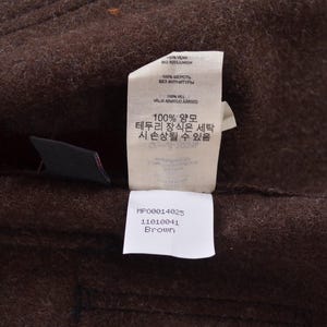 May include: A close-up shot of brown fabric with a label attached. The label contains text in multiple languages, including Korean and English, and states the fabric is 100% wool. The label also states the colour is "Brown".