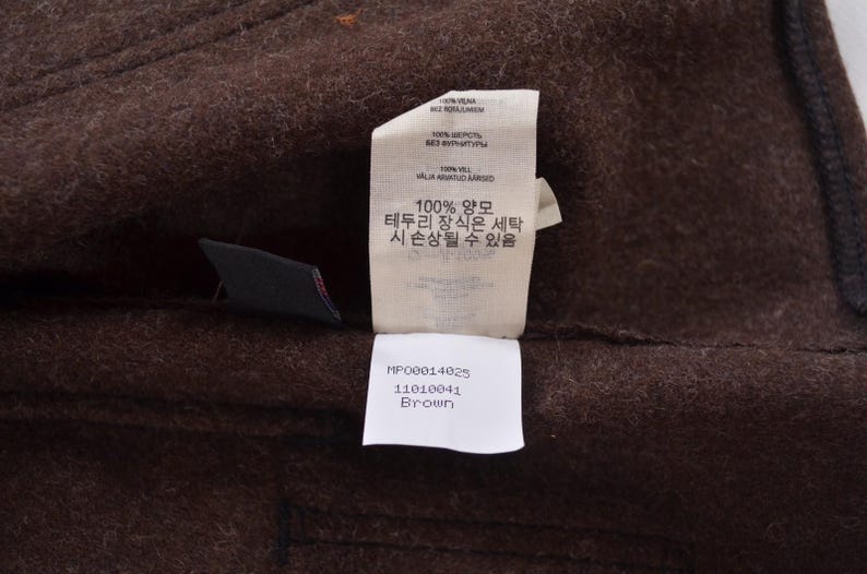 May include: A close-up shot of brown fabric with a label attached. The label contains text in multiple languages, including Korean and English, and states the fabric is 100% wool. The label also states the colour is "Brown".