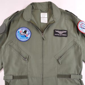 May include: Olive green flight suit with patches and a zip. Includes a "FOSTEX GARMENTS" patch and two circular patches with aviation designs. Features two zipped chest pockets and adjustable waist straps. Made of durable fabric.