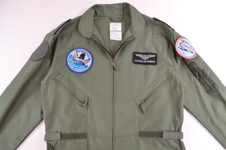 May include: Olive green flight suit with patches and a zip. Includes a "FOSTEX GARMENTS" patch and two circular patches with aviation designs. Features two zipped chest pockets and adjustable waist straps. Made of durable fabric.