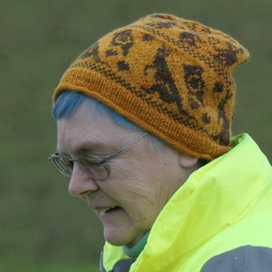May include: A woman with blue hair is wearing a brown and orange knitted hat with a geometric pattern. She is also wearing a bright yellow jacket.