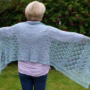 Puede incluir: Un chal de punto azul claro con un patrón de encaje. El chal está drapeado sobre los hombros de una persona de pie en un área cubierta de hierba.