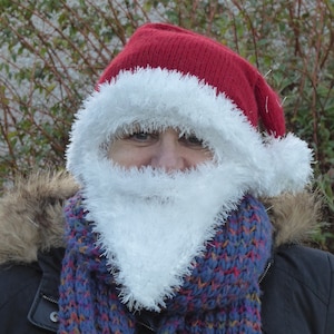 Pode incluir: Uma pessoa usando um chapéu de Papai Noel vermelho de tricô com uma borda de pele sintética branca e uma barba de pele sintética branca. A pessoa também está usando um cachecol azul e roxo e uma jaqueta preta.