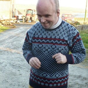 May include: A man wearing a blue and white patterned jumper with red accents. The jumper has a geometric design.