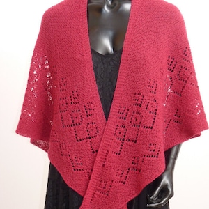 May include: A red knitted shawl with a lacy pattern. The shawl is draped over a mannequin wearing a black dress.