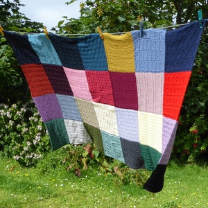May include: A colorful patchwork quilt made of knitted squares in various colors, including red, blue, green, purple, and yellow. The quilt is hanging on a clothesline outdoors.