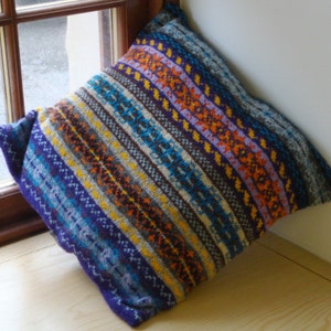 May include: A colorful knitted pillow with a striped pattern in shades of blue, purple, orange, yellow, and brown. The pillow is made of a soft, textured fabric and has a square shape.