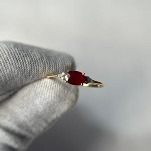Può includere: Un anello d'oro con una pietra centrale di rubino ovale e pietre d'accento bianche e nere più piccole. L'anello ha una fascia sottile e lucida. Il design è elegante e delicato. L'anello è presentato su uno sfondo neutro. Il rubino è di un rosso intenso.