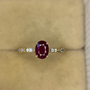 May include: A gold ring featuring an oval-cut ruby gemstone. The ring's band is adorned with small, round, clear gemstones. The ruby is a deep red color, and the ring is set against a neutral background.