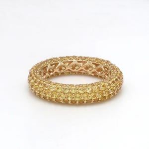 May include: A gold ring with a band covered in small, yellow gemstones. The ring has a rounded shape and is set against a white background. The gemstones are closely set together, creating a continuous surface of yellow.