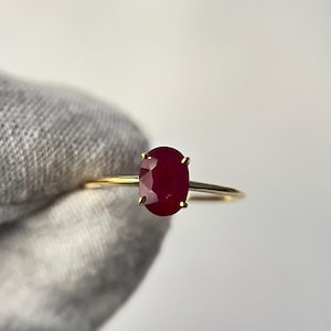 May include: A gold ring featuring a large, oval-cut ruby gemstone. The ruby is a deep, rich red color and is held in place by four delicate prongs. The band is thin and made of gold.