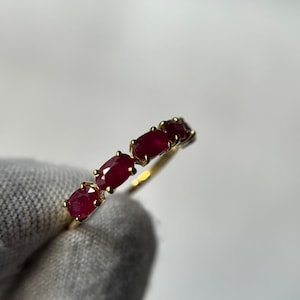 Op de afbeelding: Een gouden ring met vijf ovaal geslepen, dieprode edelstenen. De edelstenen zijn in een rij op de ringband geplaatst. De ring wordt door een gehandschoende hand tegen een witte achtergrond gehouden. Eenvoudig ontwerp.