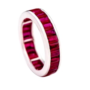 Peut inclure: Une bague en argent ornée d'une rangée continue de pierres précieuses rouges rectangulaires. Les pierres sont serties de près, créant une ligne de couleur ininterrompue. L'anneau est sur fond blanc.