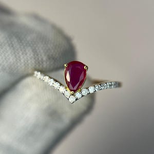 Op de afbeelding: Een gouden ring met een peervormige, dieprode robijn edelsteen. De ring heeft een V-vormig ontwerp, met kleine, heldere edelstenen langs de band. De ring wordt tegen een zachte, neutrale achtergrond gehouden.