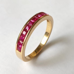 May include: A gold band ring featuring a channel setting with a row of square-cut, vibrant pinkish-red rubies. The metal has a polished, reflective finish, and the stones are set flush within the band, creating a continuous line of color.