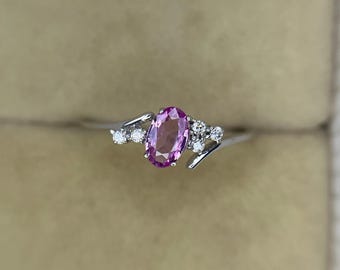 May include: A silver ring featuring an oval, pink gemstone as the centerpiece. The ring is accented with small, round, clear gemstones on either side of the main stone. The ring is set against a neutral background.
