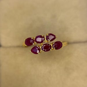 May include: A gold ring featuring a cluster of oval-cut, deep red gemstones. The gemstones are arranged in a flowing, organic design, set against a polished gold band. The ring is displayed on a neutral background.