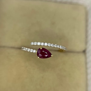 May include: A gold ring with a deep red, pear-cut gemstone as its centrepiece. The ring is embellished with a band of small, white, sparkling stones. The design is modern and elegant, suitable for special occasions.
