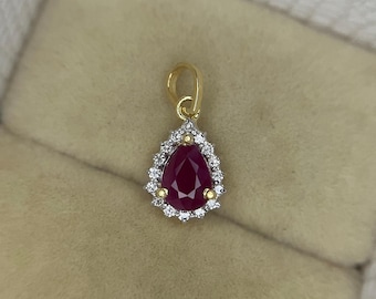 May include: A gold pendant featuring a pear-shaped, deep red gemstone surrounded by a halo of small, clear stones. The pendant has a small gold loop at the top for a chain. The setting is gold.