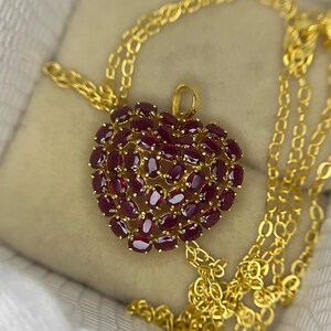 May include: A gold necklace featuring a heart-shaped pendant. The pendant is encrusted with numerous oval-cut, deep red gemstones. The necklace chain is also gold, with a delicate link design. The jewelry is presented on a soft, neutral-toned surface.