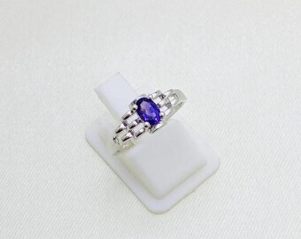 May include: A silver ring with a deep purple, oval gemstone. The ring's band has a decorative design with small rectangular accents on either side of the gemstone. The ring is displayed on a white stand against a white background.