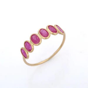 May include: A gold ring featuring six oval-cut, vibrant pink gemstones. The gemstones are set in a row along the top of the ring, creating a continuous line of color. The ring's band is a simple, polished gold.