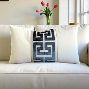 May include: A rectangular white pillow with a central blue geometric design. The pillow is on a white sofa, with a vase of red tulips in the background. The design is a repeating pattern.